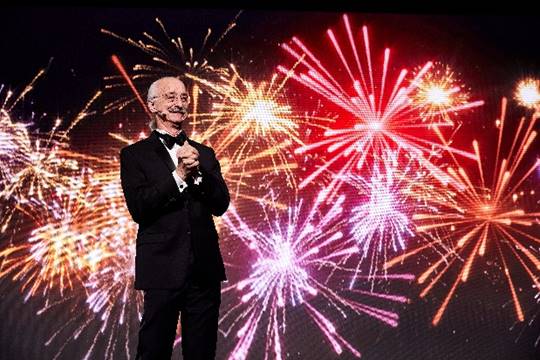 Dr. Woodie Flowers, dressed in a tuxedo, smiles and clasps his hands together. Fireworks explode in the background.