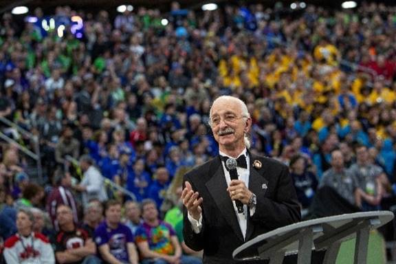 Dr. Woodie Flowers, dressed in a tuxedo, is holds a microphone and addresses a large crowd that surrounds him.